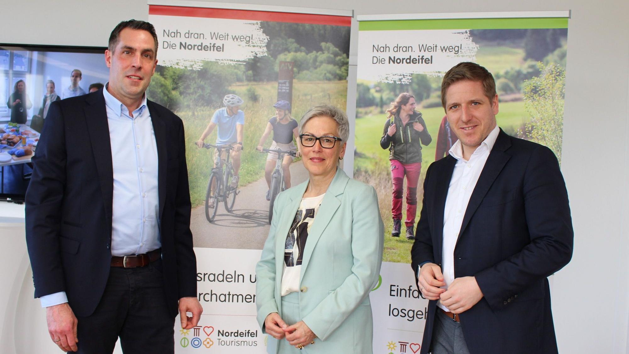 Zwei Männer und eine Frau stehen vor Roll-ups mit dem Slogan „Nah dran. Weit weg! Die Nordeifel“.
