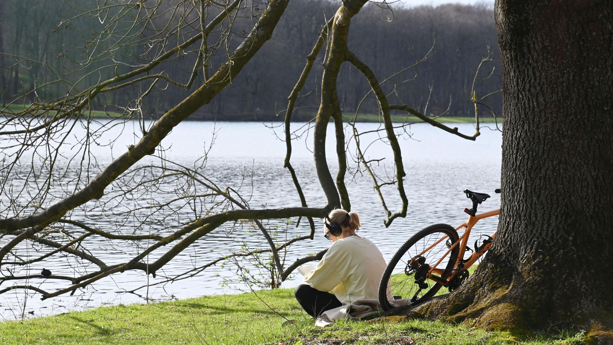 Eine Frau sitzt am Decksteiner Weiher in Köln neben ihrem Fahrrad und hört bei sommerlichen Temperaturen Musik.