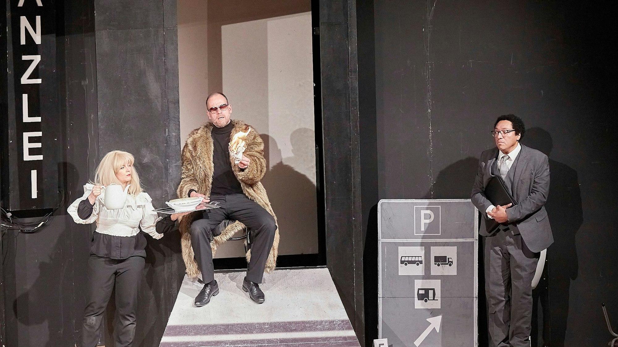 Eine Frau und zwei Männer, einer im Pelzmantel, der andere im Anzug, stehen auf der Bühne.