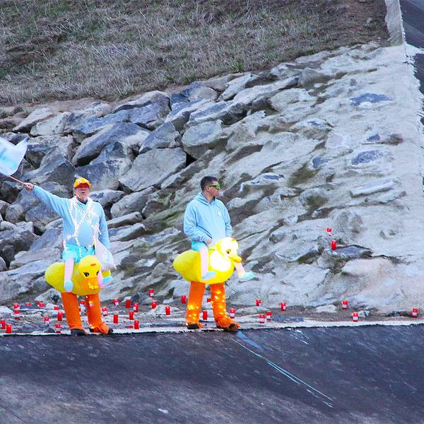 Zwei Männer, die gelbe Entchen-Kostüme umgebunden haben, stehen an der Steinbach. Einer schwenkt eine hellblaue Fahne.