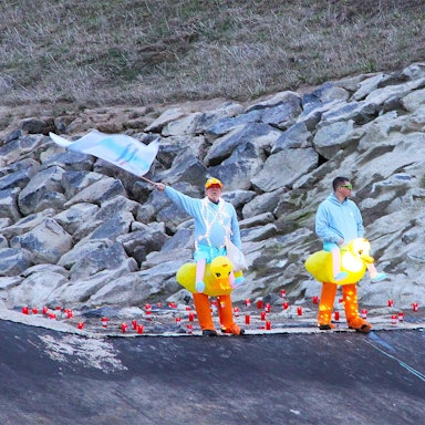 Zwei Männer, die gelbe Entchen-Kostüme umgebunden haben, stehen an der Steinbach. Einer schwenkt eine hellblaue Fahne.