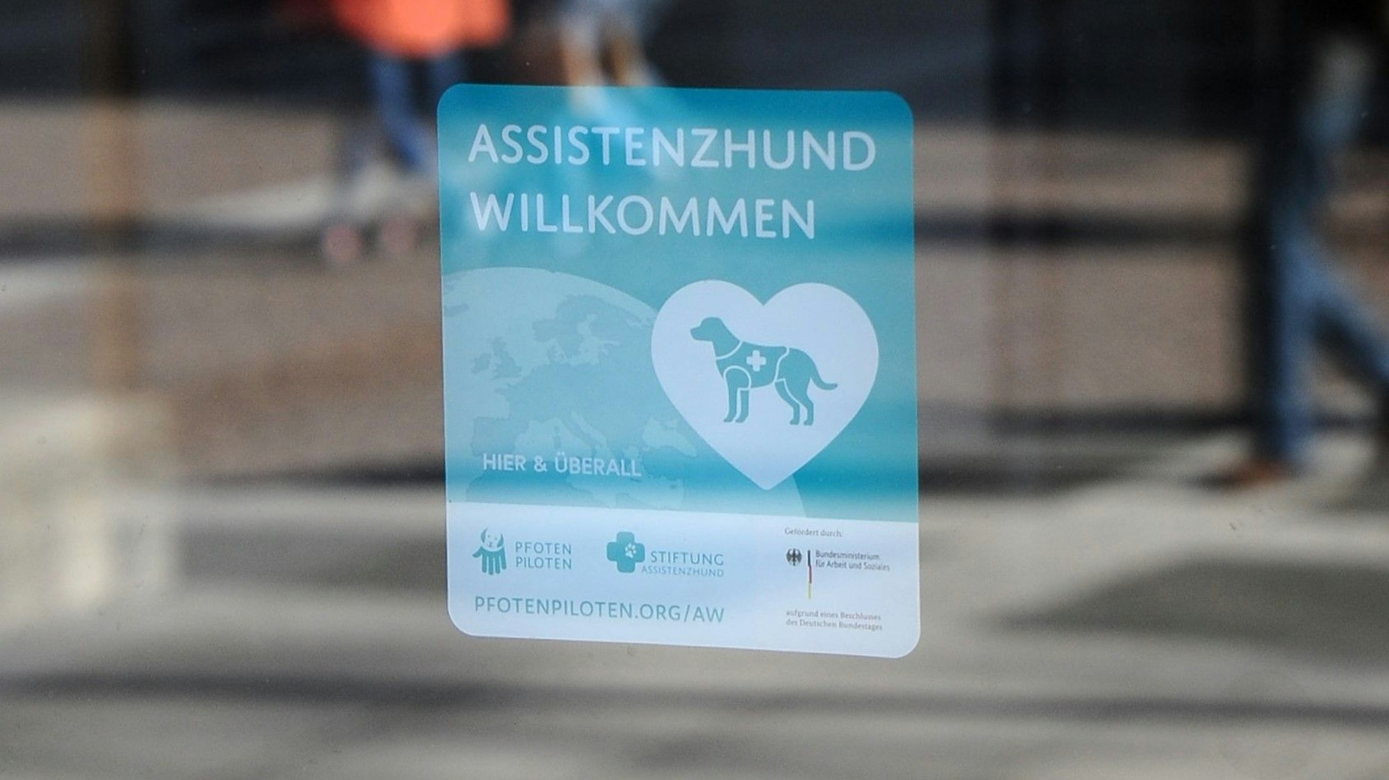 Auf einem Aufkleber ist der Schriftzug „Assistenzhund willkommen“ zu sehen, darunter ist ein Assistenzhund in einem Herz abgebildet.