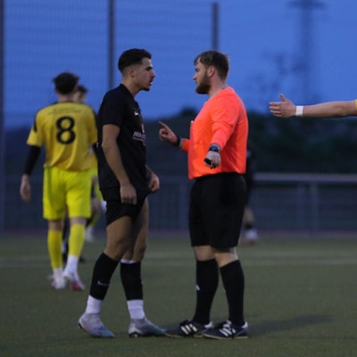 Schiedsrichter Eric Strohmeier spricht auf dem Fußballplatz mit dem JSG-Spieler Arber Jashanica, ein Erftstädter Akteur beobachtet die Szene mit ausgebreiteten Armen.