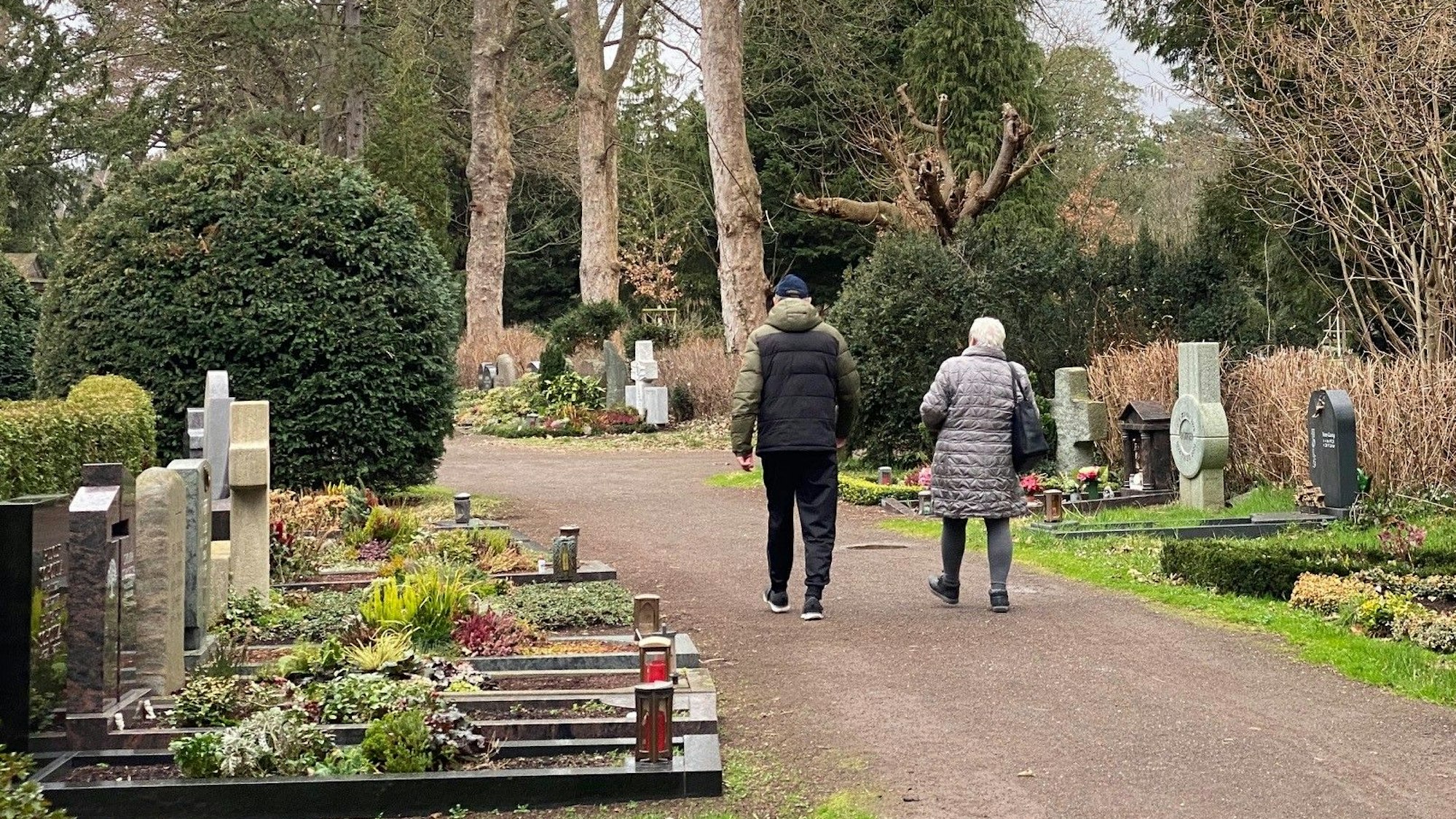 Zu sehen ist der Brühler Südfriedhof mit Grabstätten und zwei Besuchern.