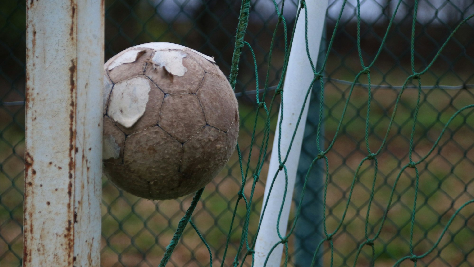Das Bild zeigt einen alten Fußball, der zwischen Tor und Zaun eingeklemmt ist.