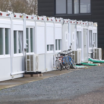 Wohncontainer auf einem Schotterplatz. Vor den Containern stehen Lüftungsgeräte, an einige sind Fahrräder angelehnt.