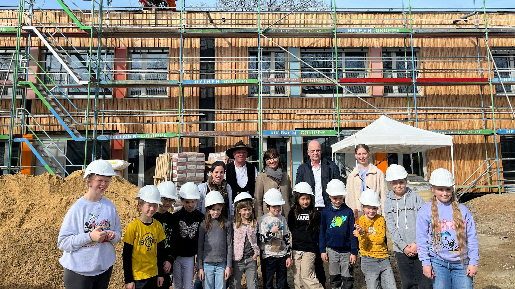 Schülerinnen und Schüler mit Bauhelmen und einige Erwachsene stehen vor einem Gebäude mit einem Baugerüst.