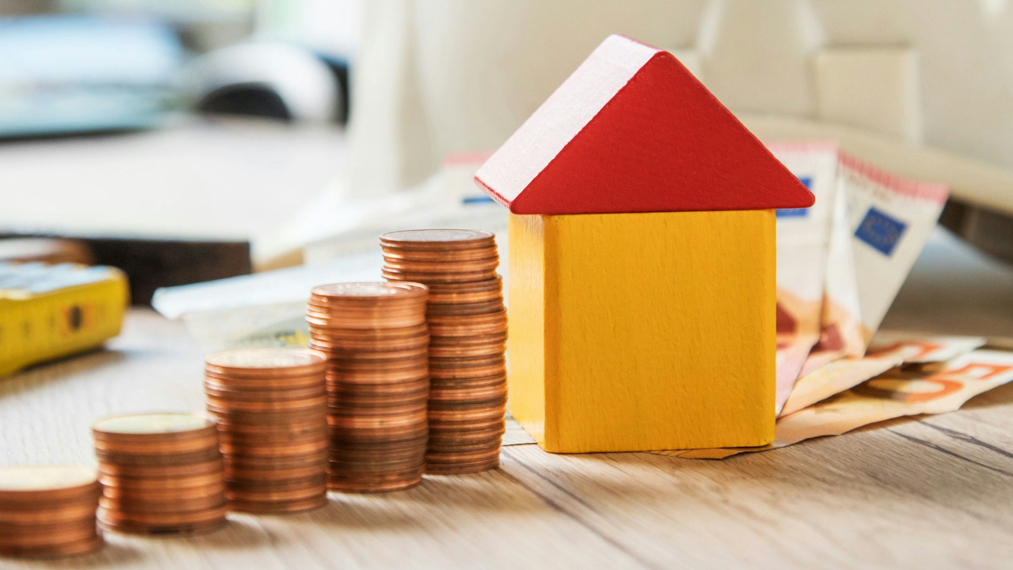 Ein Haus aus Holzklötzen steht neben aufgestapeltem Kleingeld als Symbolbild für die Grundsteuer.