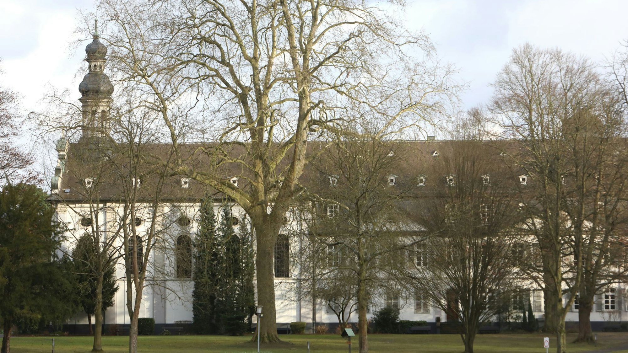 Ein weißes, historisches Gebäude mit einem kleinen Kirchturm steht hinter hohen Bäumen.