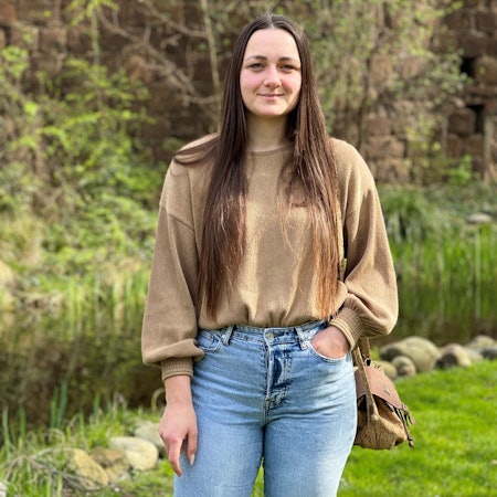 Eine junge Frau mit langen, dunklen Haaren steht vor grünen Pflanzen.