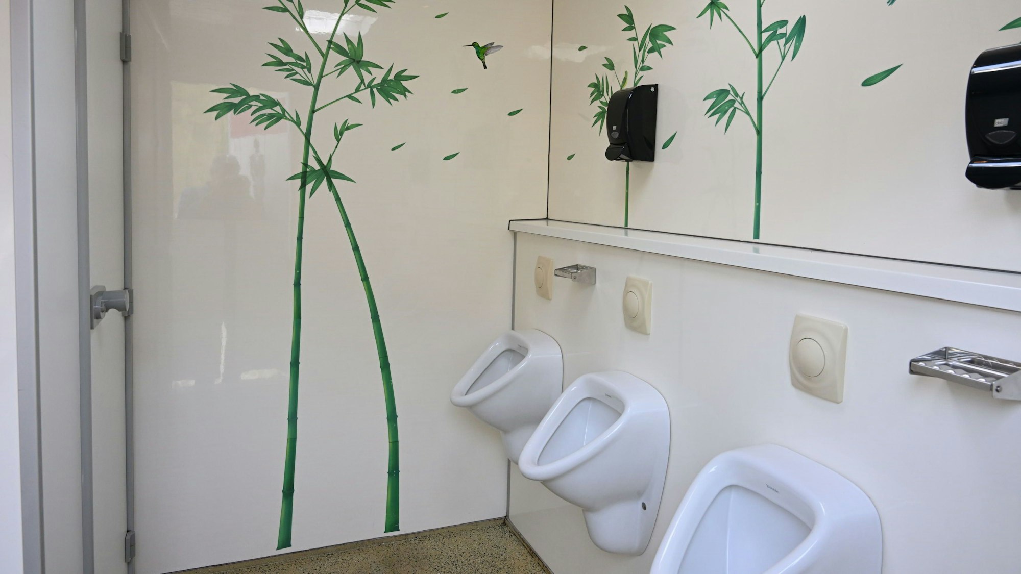 Das Innere eines Toilettenwagens: drei weiße Urinale, weiß lackierte Wände mit Palmenmotiven darauf.