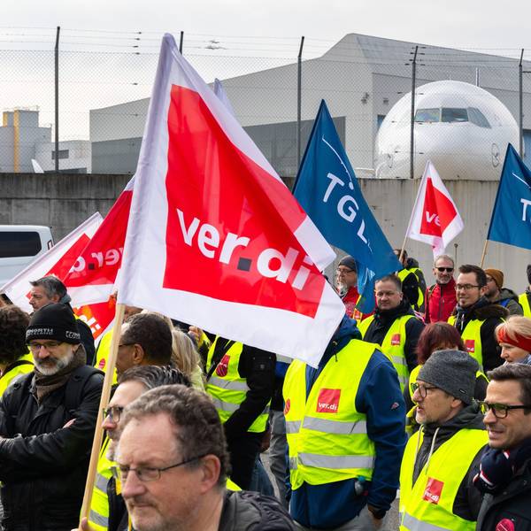 Ein Protestzug von Streikenden zieht mit Bannern und Verdi-Fahnen vom Lufthansa Aviation Center zum Terminal 1. Im Hintergrund ist ein Flugzeug der Lufthansa zu sehen.