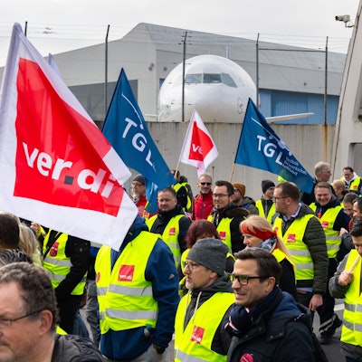 Ein Protestzug von Streikenden zieht mit Bannern und Verdi-Fahnen vom Lufthansa Aviation Center zum Terminal 1. Im Hintergrund ist ein Flugzeug der Lufthansa zu sehen.