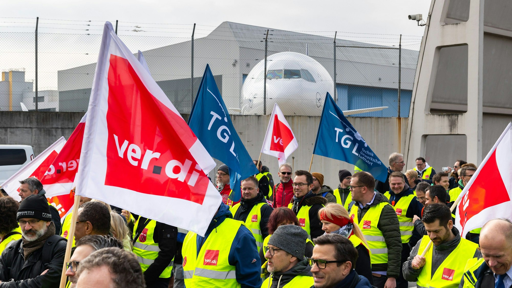 Ein Protestzug von Streikenden zieht mit Bannern und Verdi-Fahnen vom Lufthansa Aviation Center zum Terminal 1. Im Hintergrund ist ein Flugzeug der Lufthansa zu sehen.