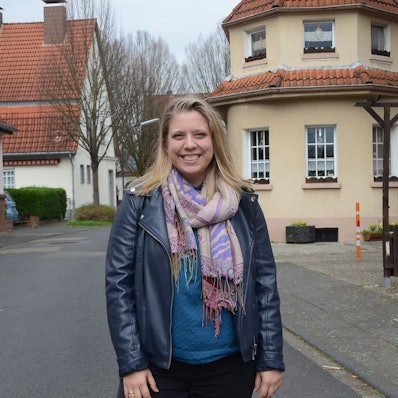 Eine junge Frau mit blonden Haaren auf einer Straße. Hinter ihr sind ältere Häuser einer Siedlung zu sehen.