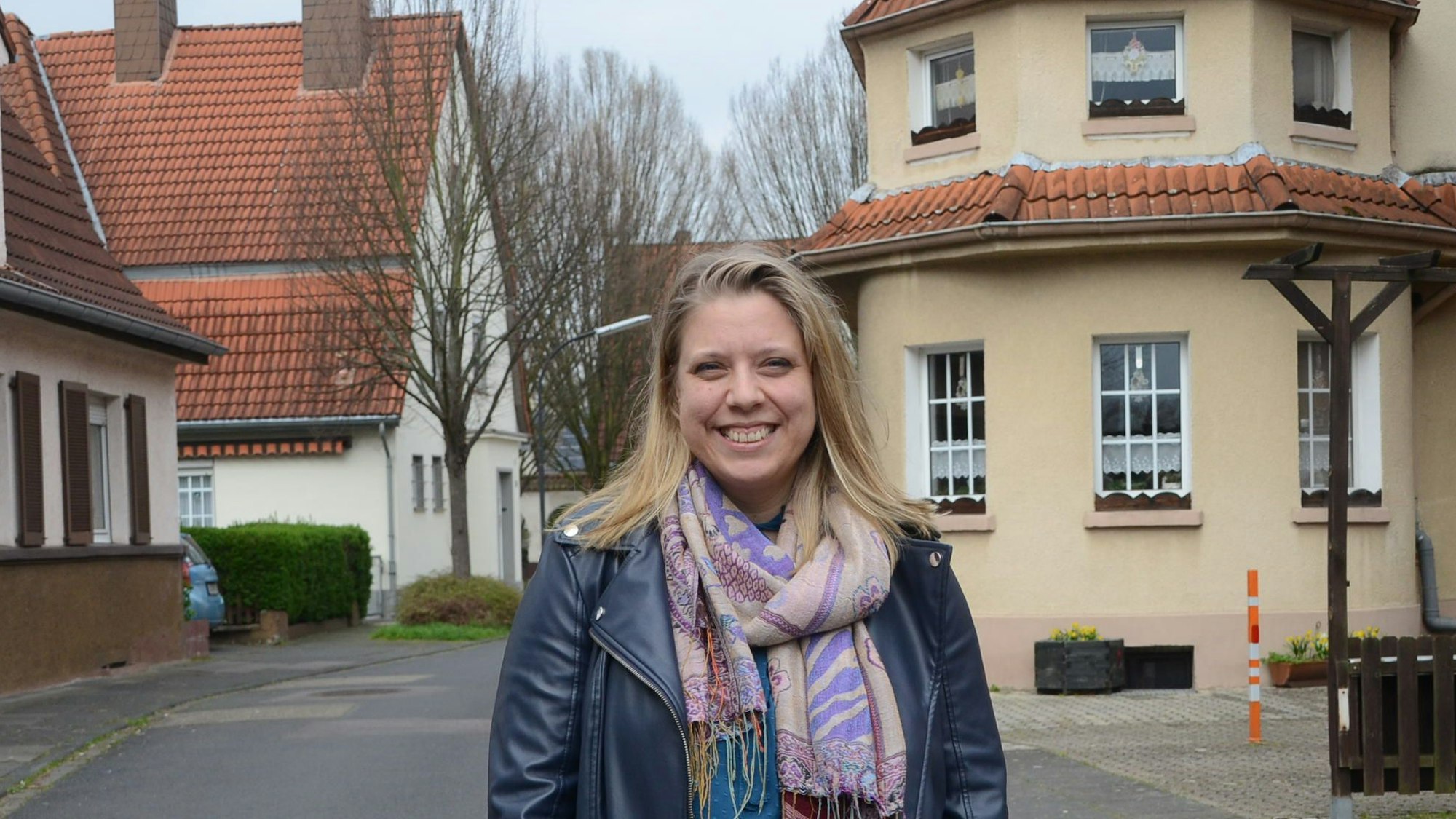 Eine junge Frau mit blonden Haaren auf einer Straße. Hinter ihr sind ältere Häuser einer Siedlung zu sehen.