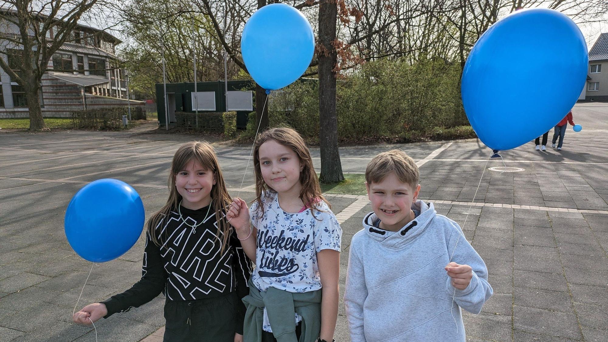 Drei Schüler und Schülerinnen stehen mit je einem Ballon in der Hand auf dem Schulhof.
