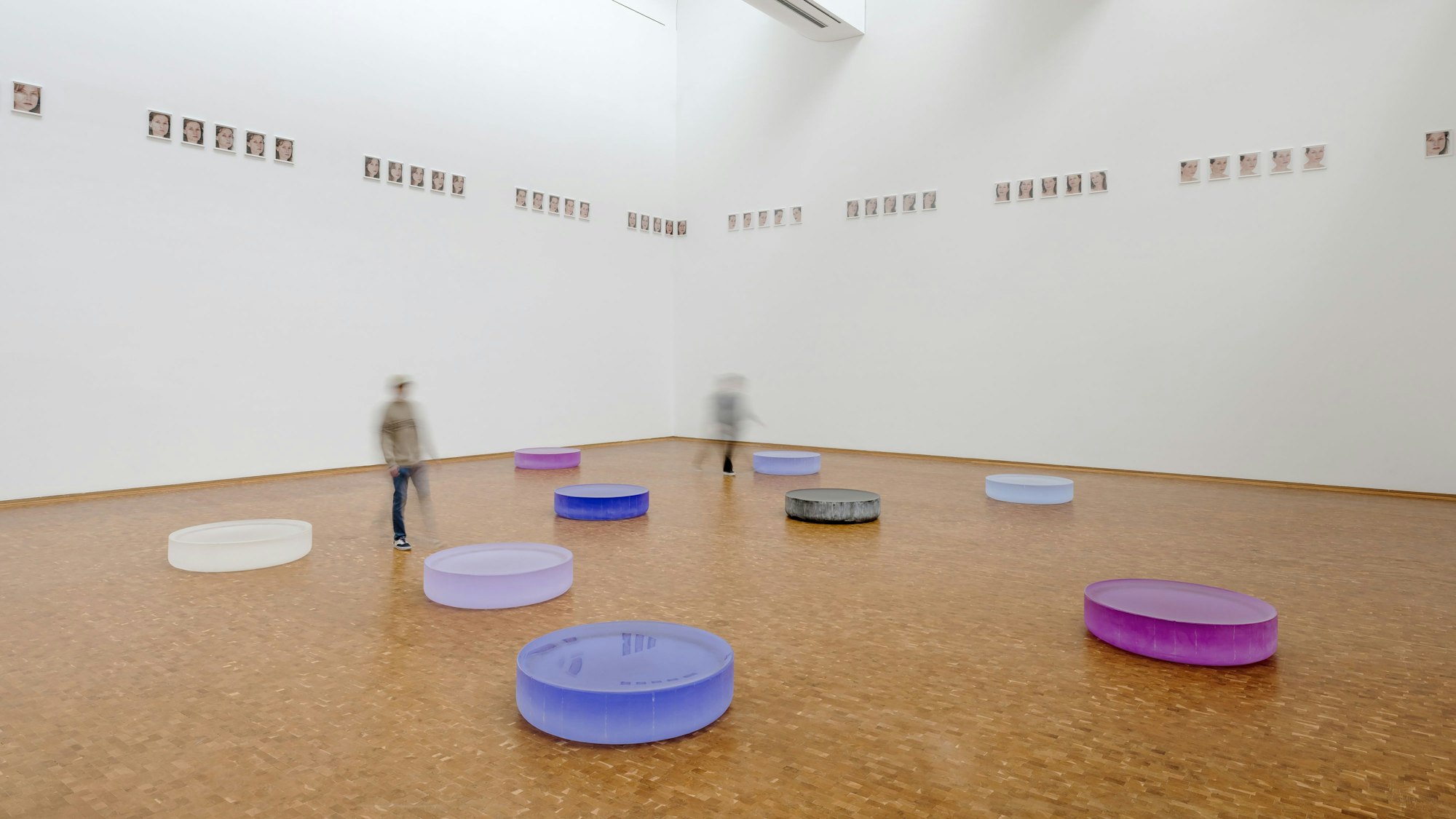 Tröge aus Glas wirken wie mit Wasser gefüllt. Weit oben hängen Porträts von Isabelle Huppert.