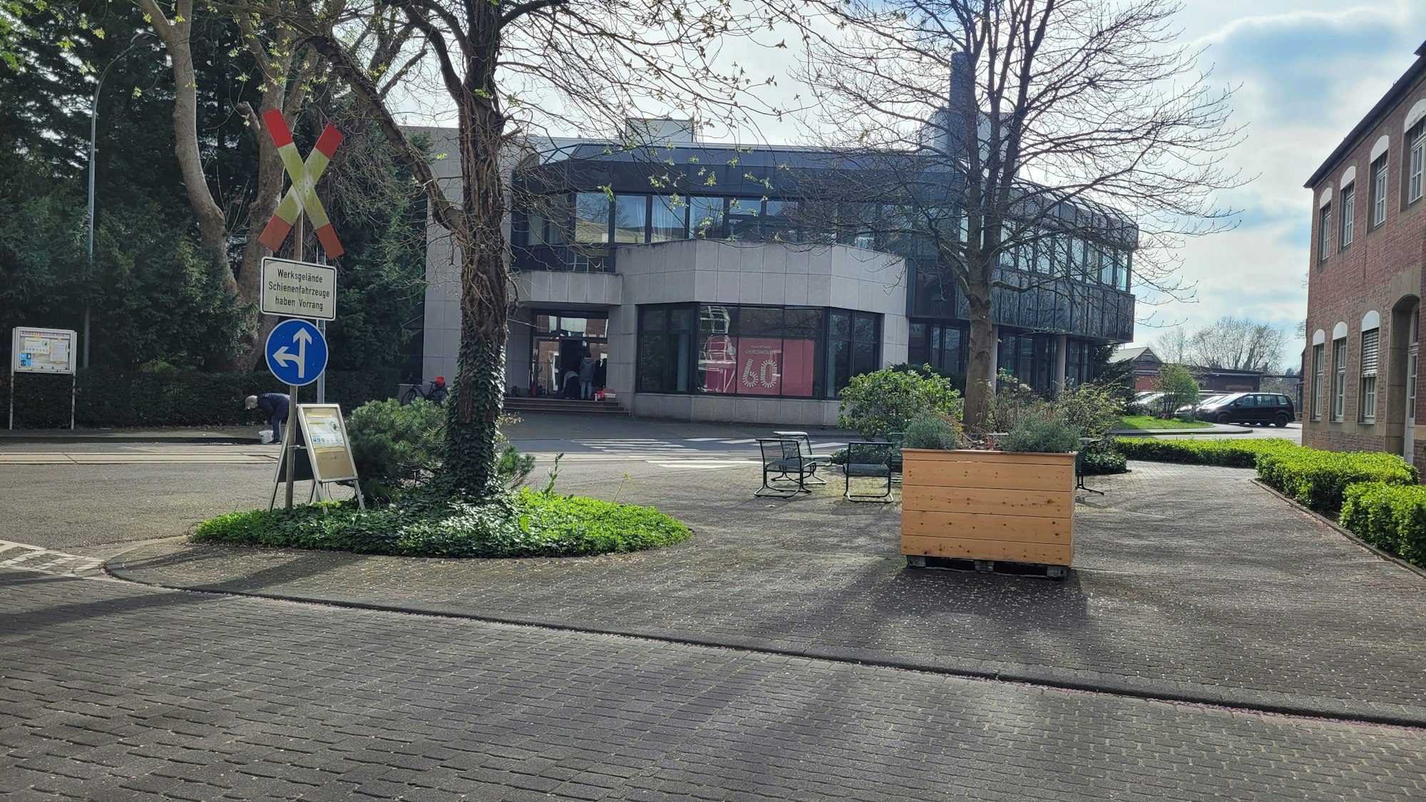 Das eckige zweigeschossige Gebäude ist zu sehen, mit Bäumen und Bänken im Vordergrund.