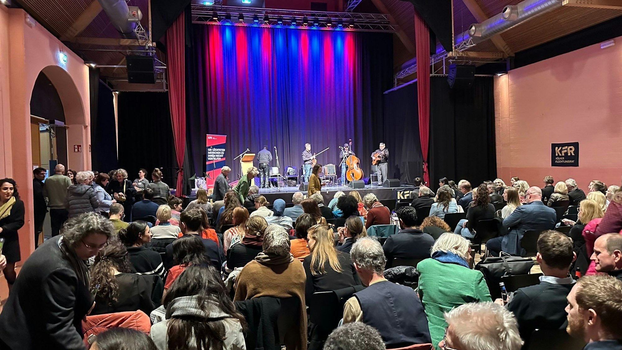 In einem bestuhlten Raum sitzen Leute und Musiker stehen auf einer Bühne.