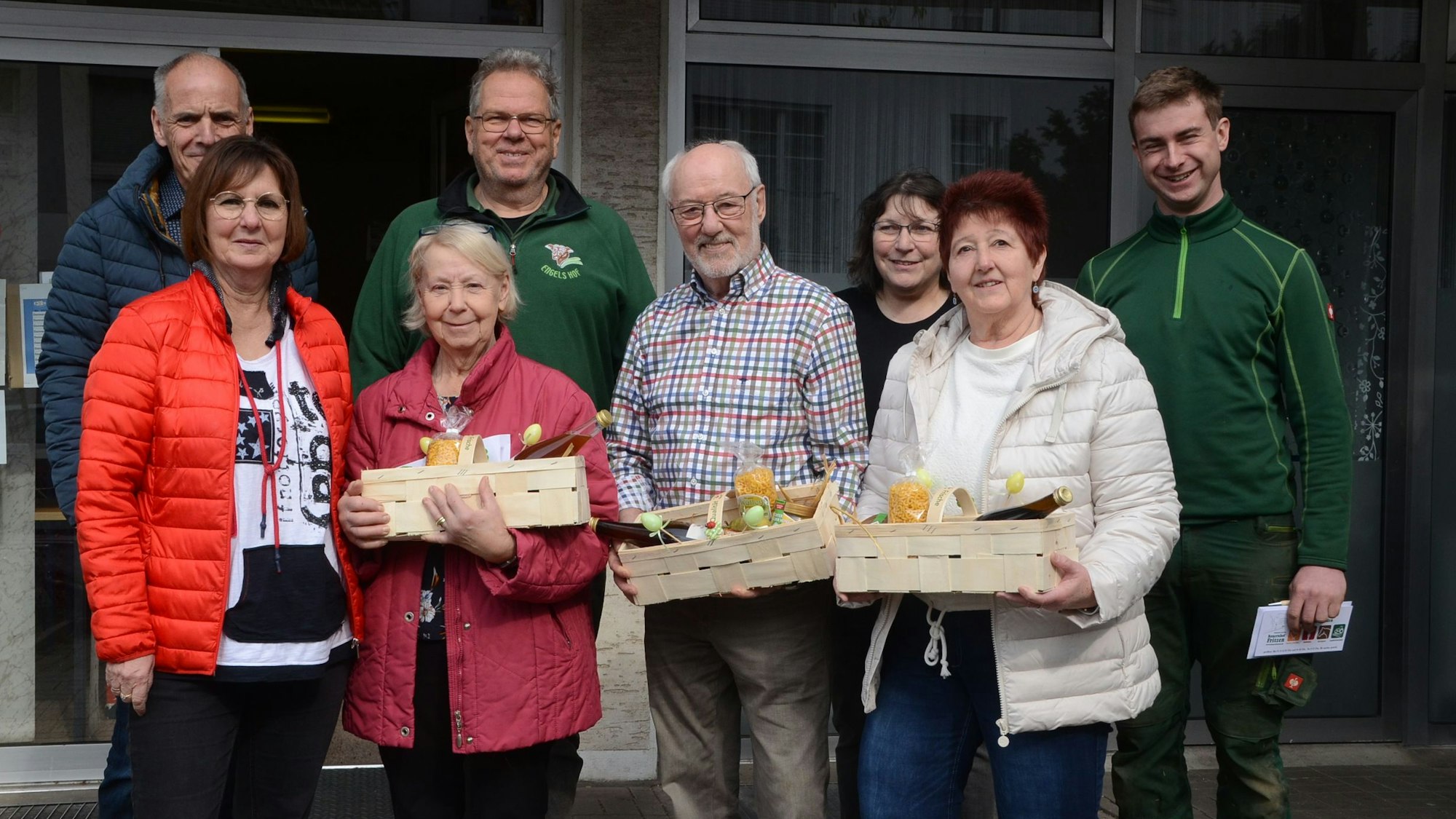 Männer und Frauen stehen in einer Gruppe zusammen. Drei halten einen Spankorb mit Gemüse, Obst und einem Honigglas.