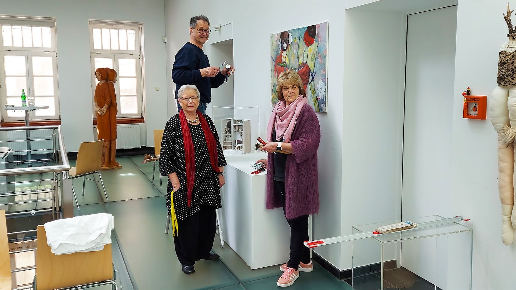 Das Bild zeigt einen Mann, der auf einem Stuhl steht, um etwas anzubringen, und zwei Frauen, die etwas in der Hand halten.