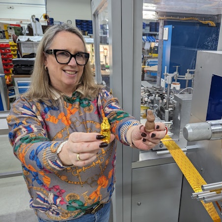 Eine Frau steht vor einer großen Maschine und zeigt jeweils einen Schokohasen mit und ohne Einwicklung.