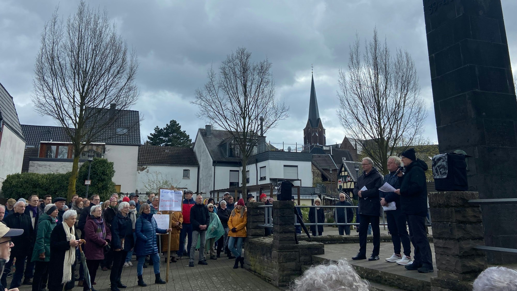 Menschen stehen auf dem Zündorfer Marktplatz vor dem Kriegerdenkmal.