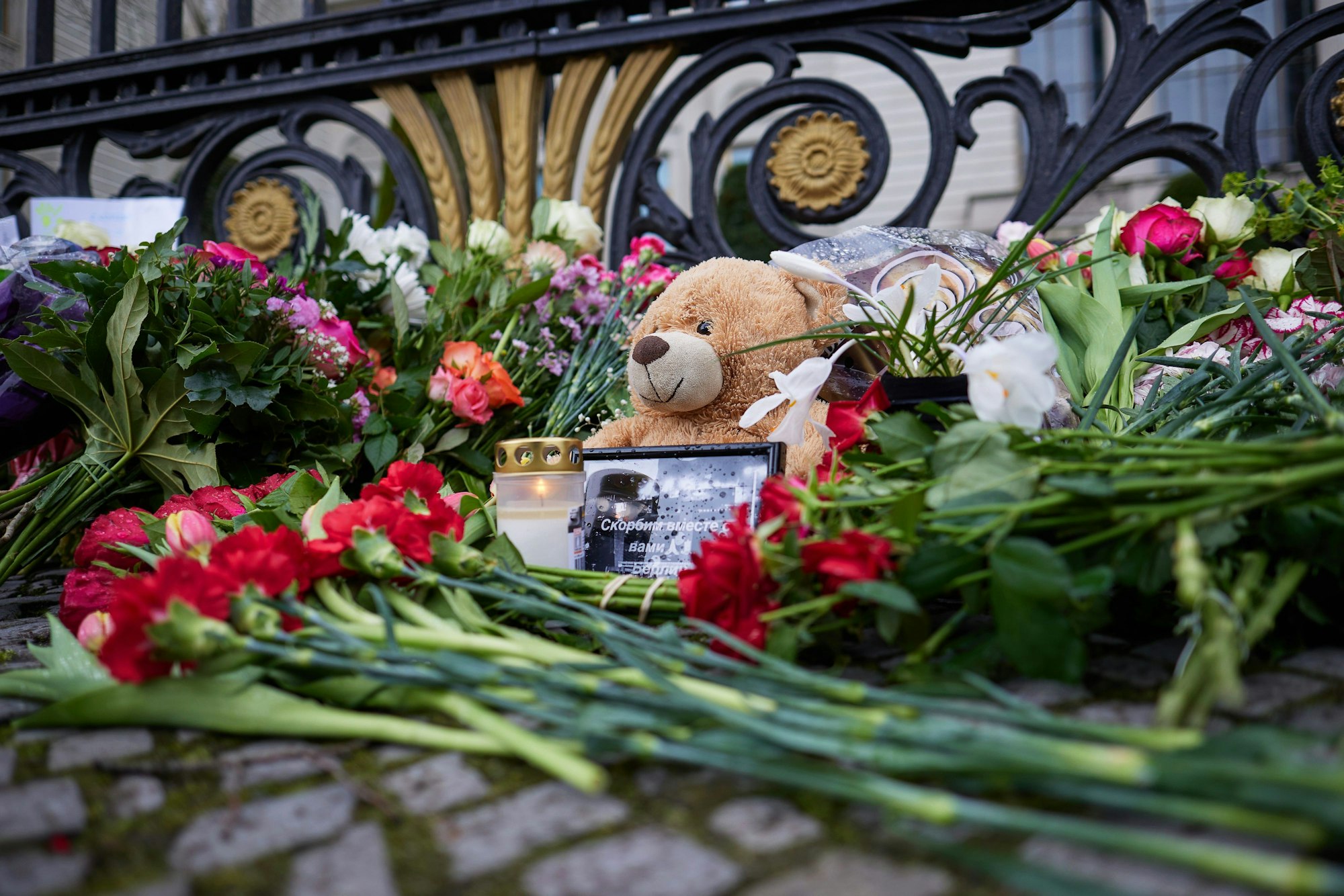 Eine Nahaufnahme zeigt einen Teddybären mit einem Bild und einer Kerze, umgeben von Blumen.