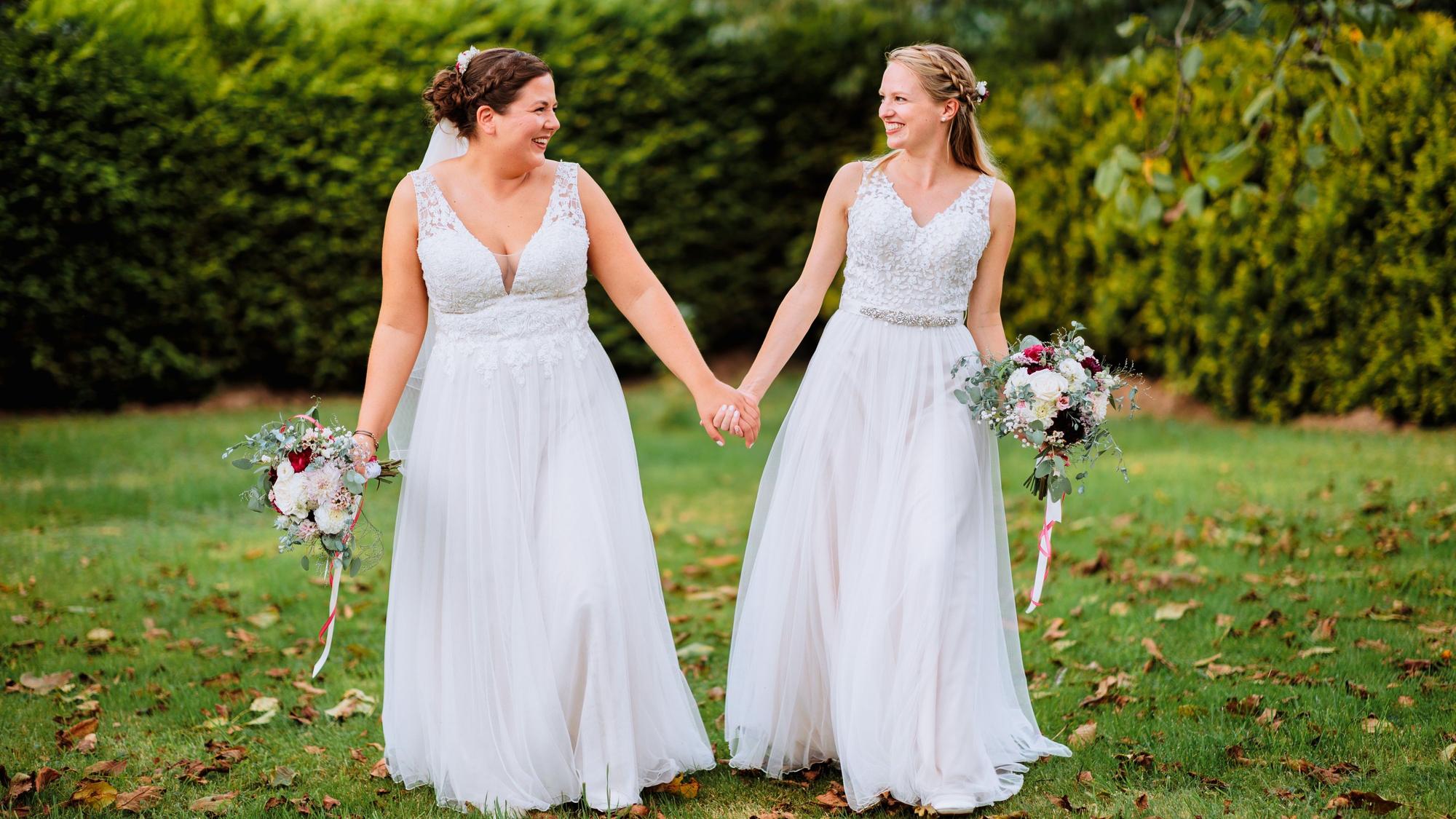 Laura und Christel Wendeler tragen ihre Brautkleider und schauen sich an.