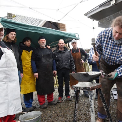 Ein Schmied in einem Museum bei der Arbeit. Besucher schauen ihm zu.