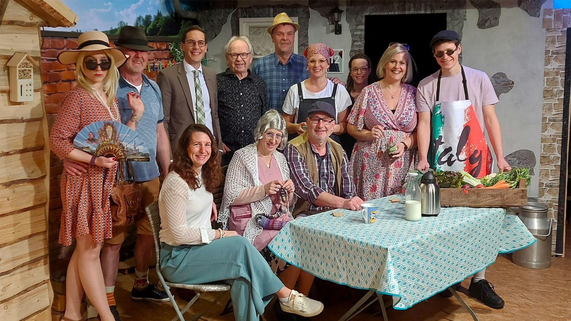 Die Schauspieler sitzen und stehen hinter einem Tisch mit Requisiten.