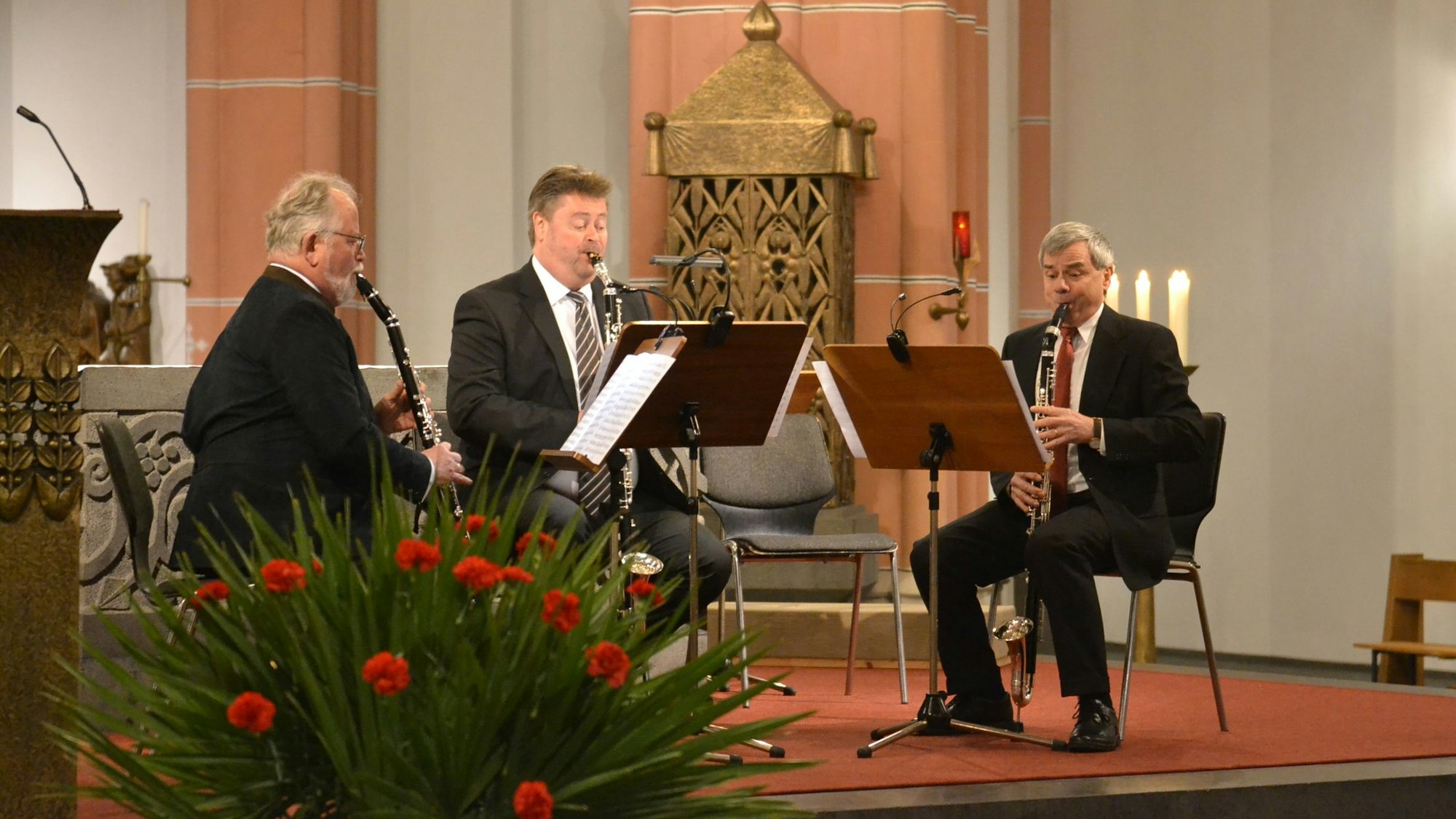 Drei Instrumentalisten musizieren vor dem Altar in der Euskirchener Herz-Jesu-Kirche. Im Vordergrund Blumen, im Hintergrund Kerzen.