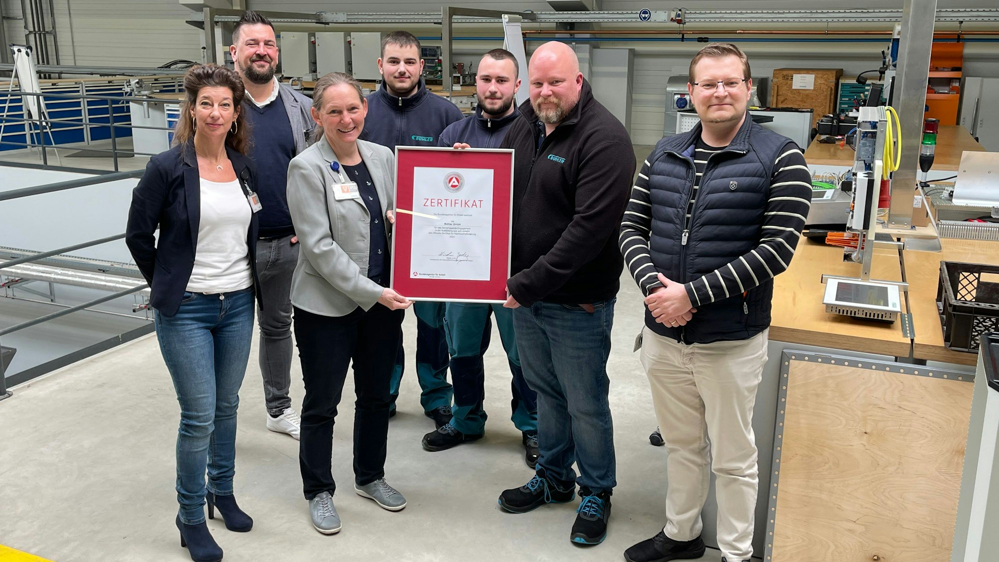 Eine Gruppe Männer und Frauen steht mit einem Bilderrahmen und dem Ausbildungszertifikat in einer Produktionshalle eines Unternehmens.