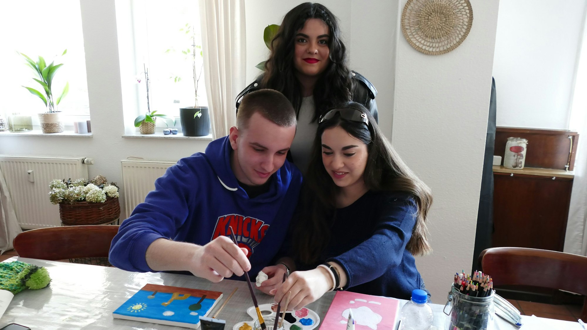 Eine junge Frau und ein Mann tauchen ihre Pinsel in eine Farbpalette, eine andere junge Frau steht hinter ihnen.