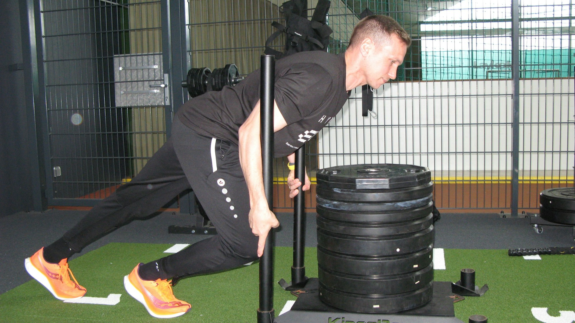 Der 38-jährige Roman Hopp, athletische Figur, schwarzes T-Shirt, schwarze Trainingshose, stemmt sich gegen einen mit großen Hantelscheiben beladenen Schlitten aus einer Metallplatte und zwei senkrecht eingelassen Stangen.