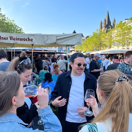 Eine Menschenmenge steht auf dem Heumarkt. Personen trinken Wein.
