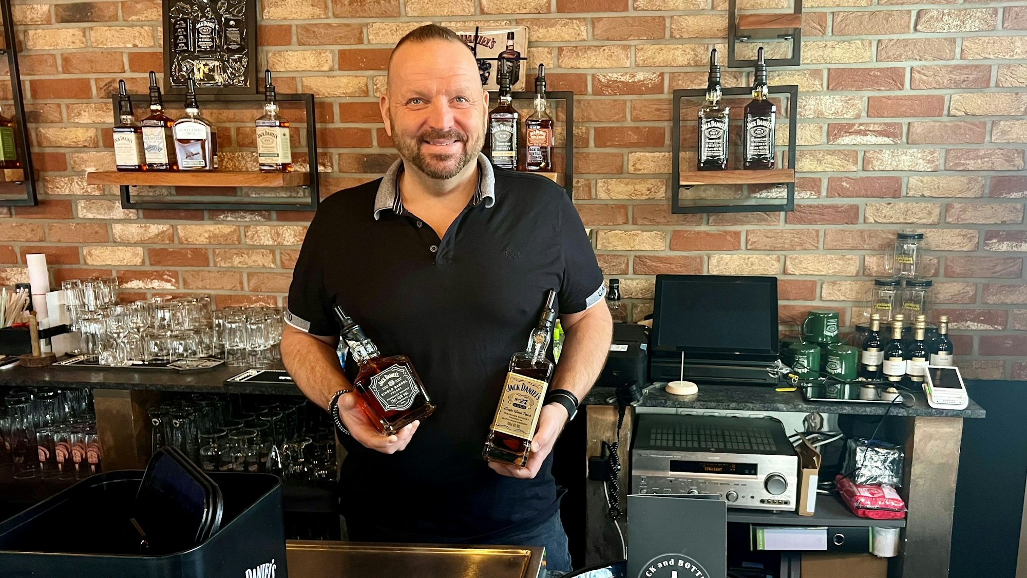 Ein Kölner Wirt hinter dem Tresen mit zwei Whiskey-Flaschen im Arm