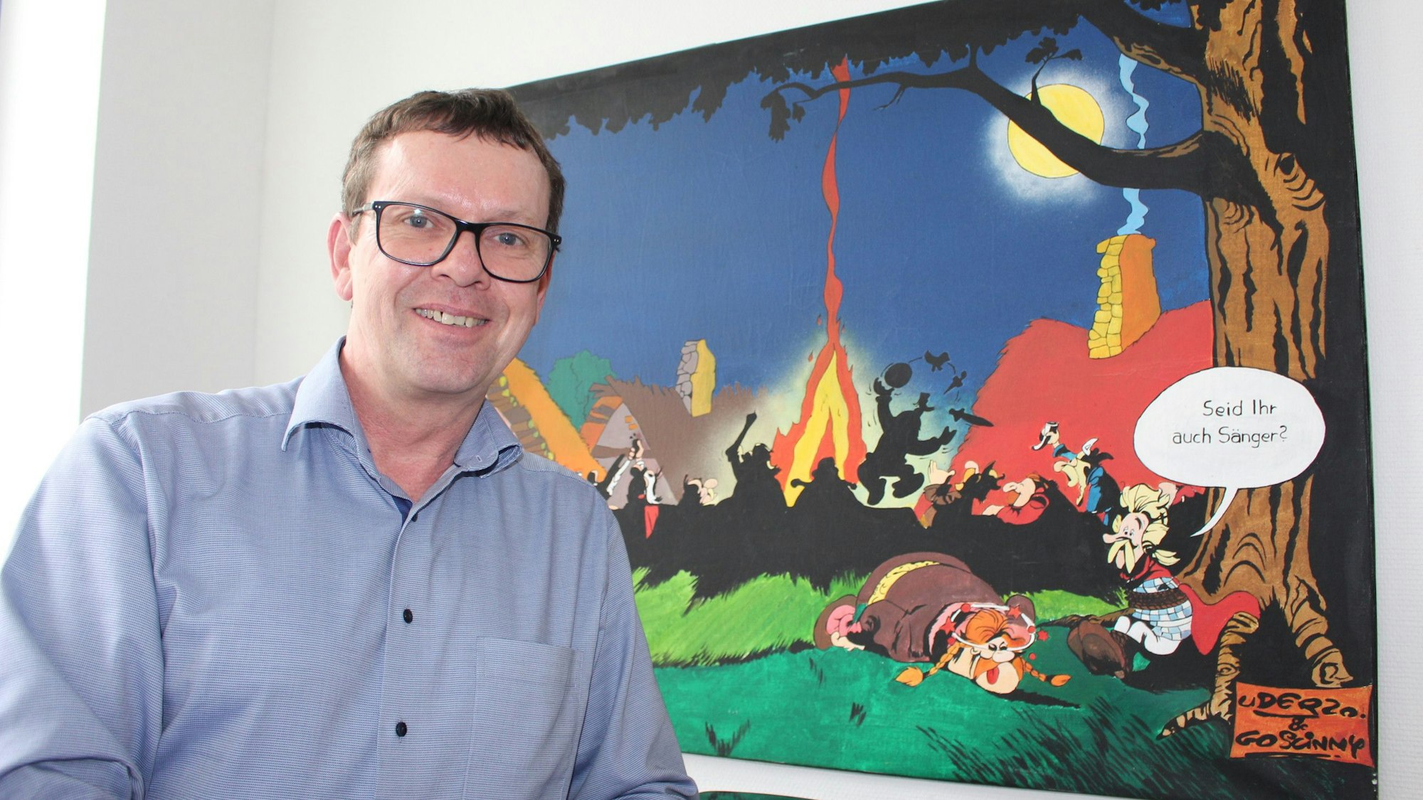 Erdmann Bierdel vor einem Bild mit einer Szene aus einem Asterix-Comic. In einer Sprechblase steht: „Seid Ihr auch Sänger?“ Das Bild haben ihm Kollegen geschenkt, nachdem er eine selbst getextete „Jugendamtsleiterballade“ vorgetragen hatte.