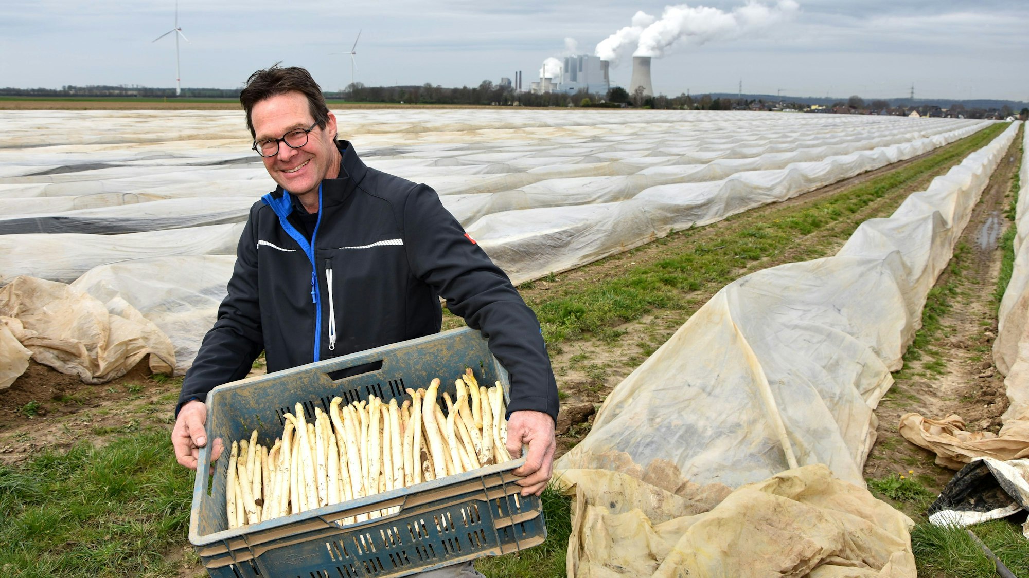 Ein Mann trägt eine Kiste Spargel von einem Feld, im Hintergrund steht ein Kraftwerk.