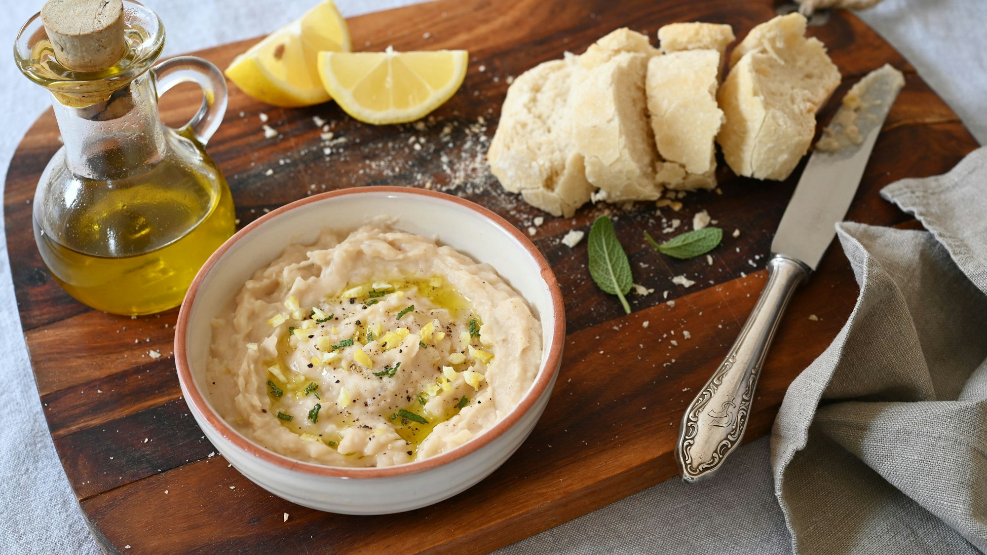 Auf einem Holzbrett steht eine Schüssel mit den pürierten Bohnen, Öl, Zitronen und Baguettescheiben.