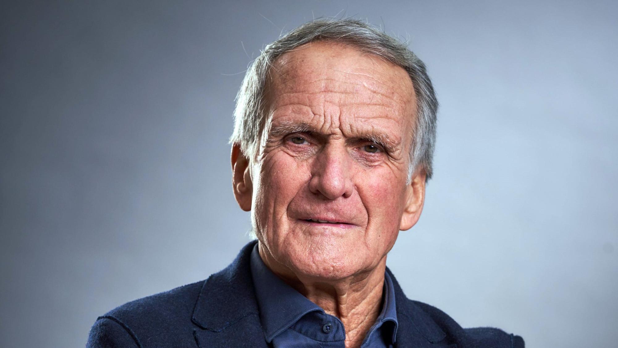 Wolfgang Overath schaut mit verschränkten Armen und gewohnt verkniffenem Blick in die Kamera.