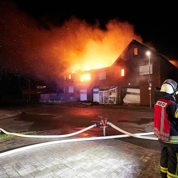 Drei Feuerwehrleute stehen vor einem Haus in Schleiden-Herhahn, das in Vollbrand steht.