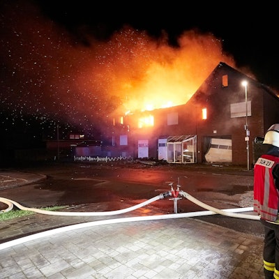 Mehrere Feuerwehrleute stehen vor einem brennenden Haus.