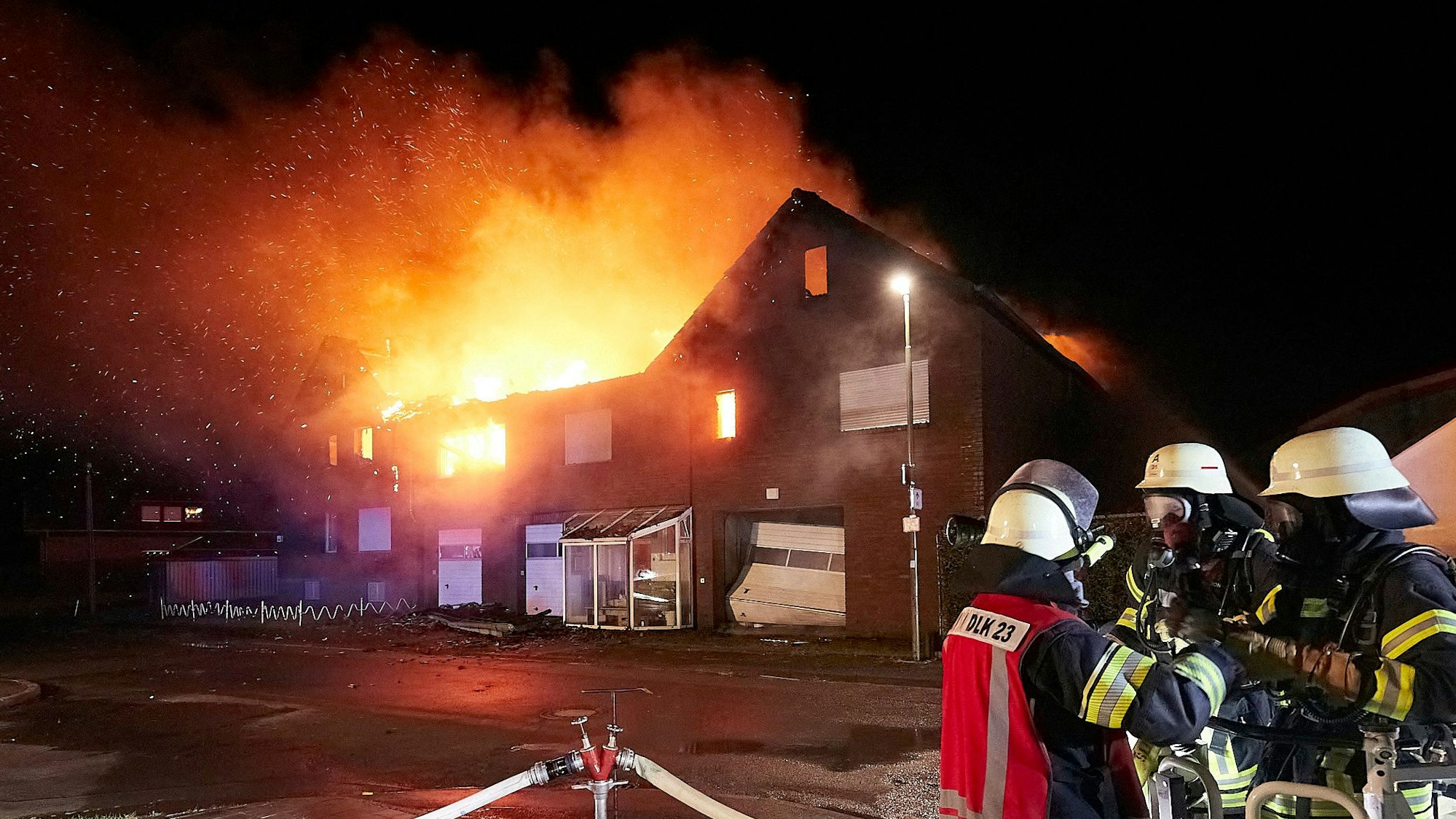Drei Feuerwehrleute stehen vor einem Haus in Schleiden-Herhahn, das in Vollbrand steht.