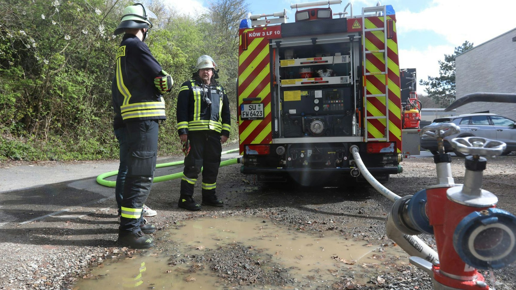 Zwei Feuerwehrleute stehen hinter einem Löschfahrzeug, an einem Hydranten ist ein Löschschlauch angebracht.
