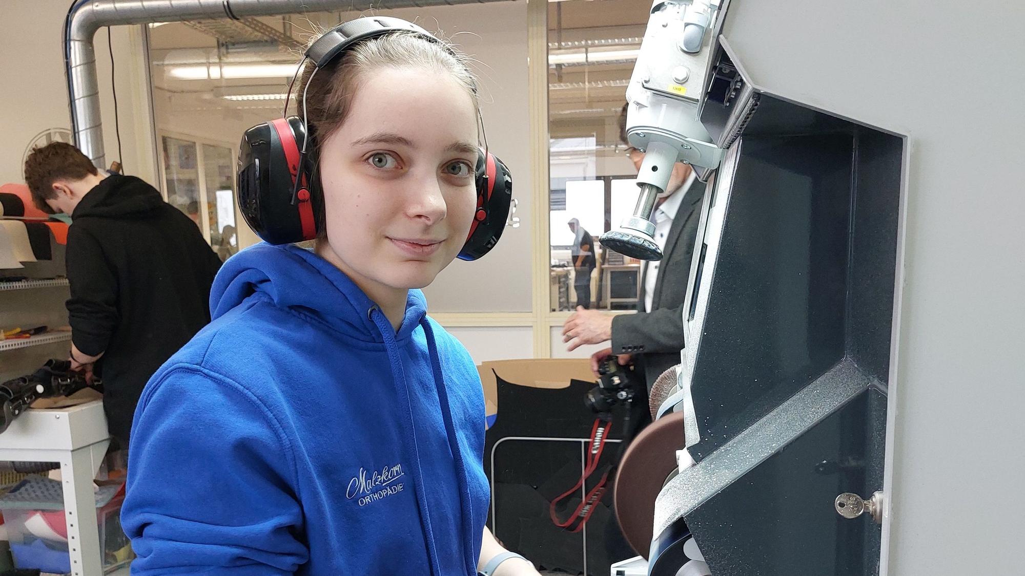 Eine junge Frau in einem Pullover steht mit Ohrschützern an einer Maschine.