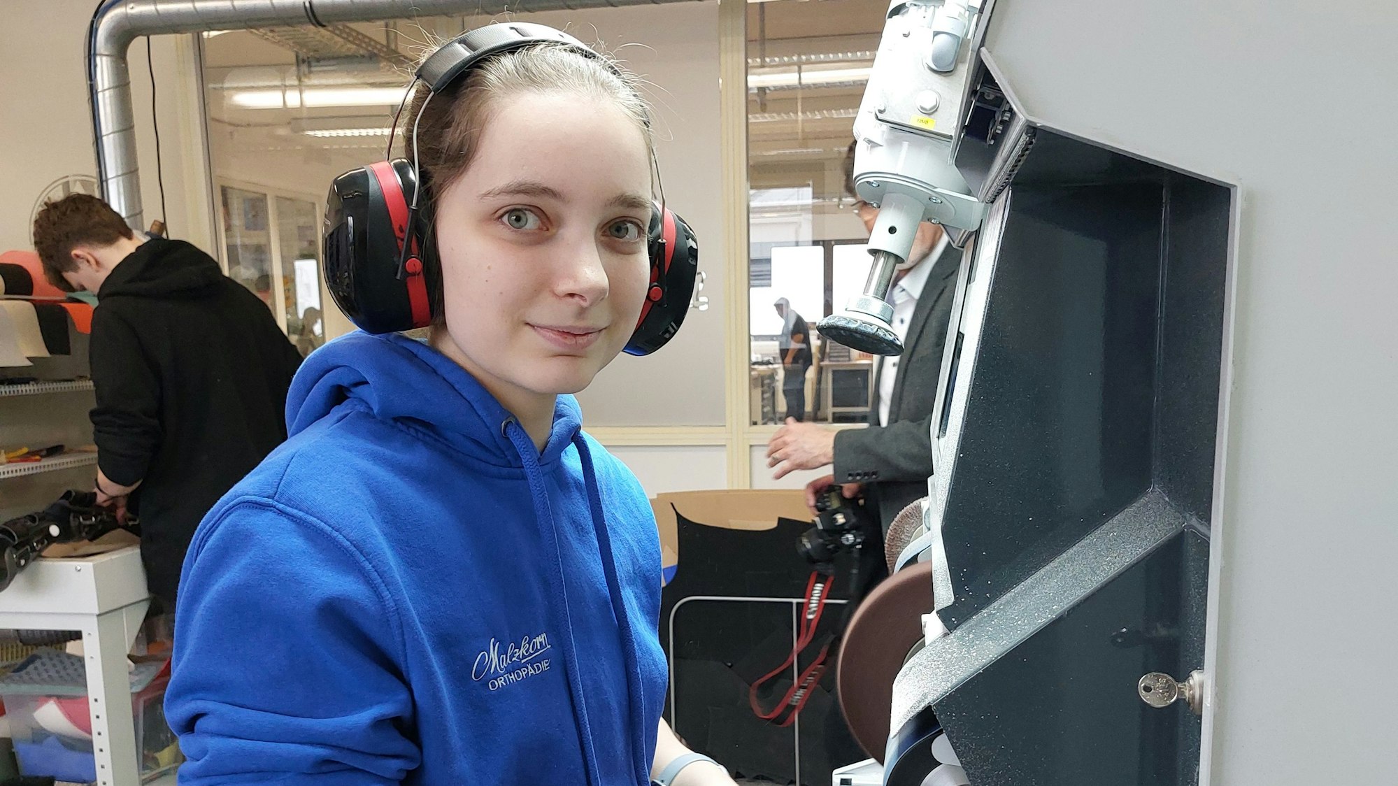 Eine junge Frau in einem Pullover steht mit Ohrschützern an einer Maschine.