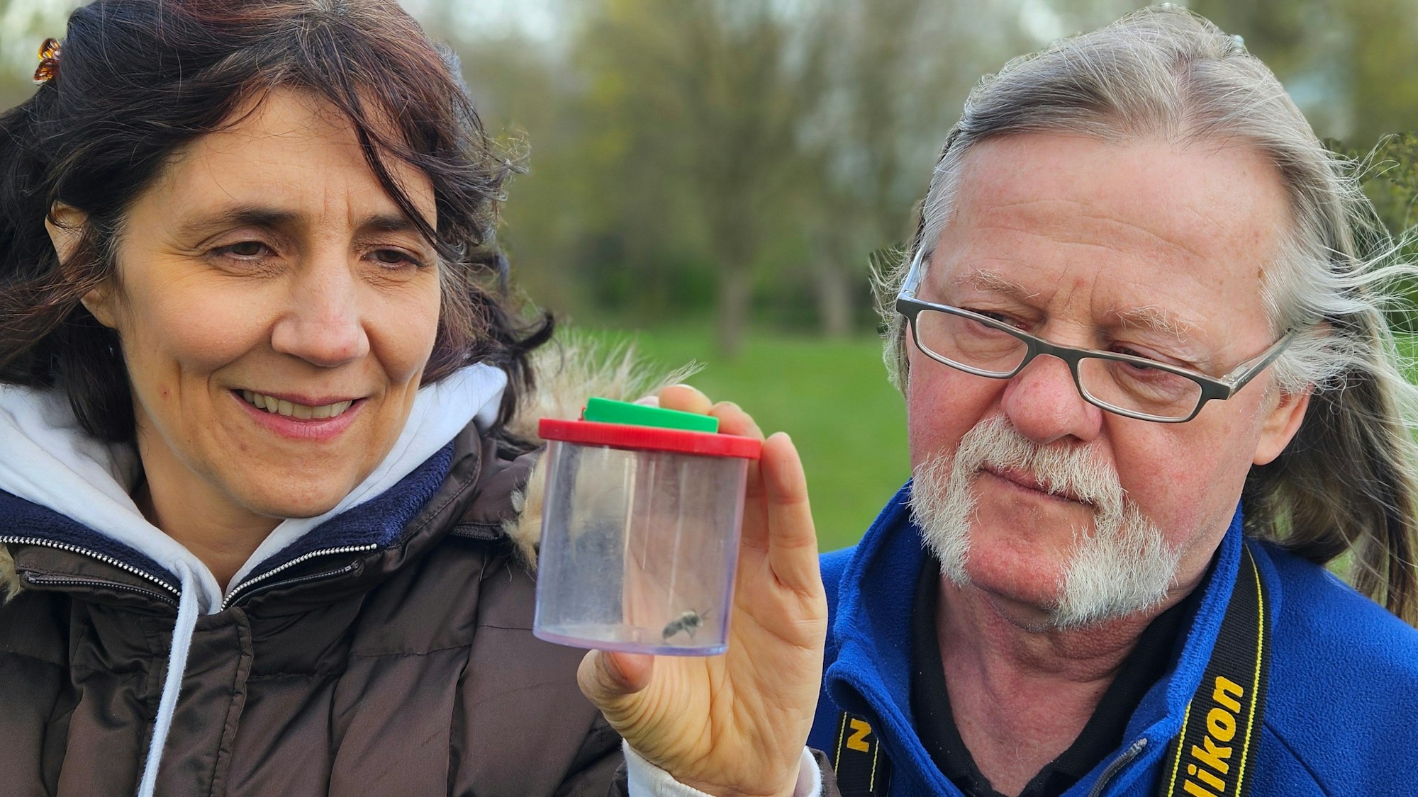 Eine Frau und ein Mann betrachten eine Biene in einer Becherlupe.