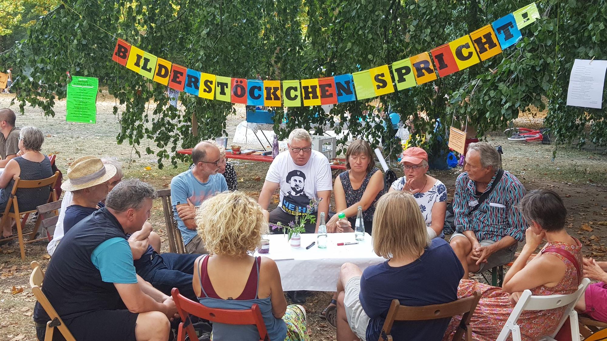 Menschen sitzen im Spätsommer an einem Tisch und diskutieren bei der sechsten Auflage der „Speakers' Corner“ im Blücherpark. Im Hintergrund ist ein Banner mit der Aufschrift „Bilderstöckchen spricht“ zu sehen. Foto von Bernd Schöneck